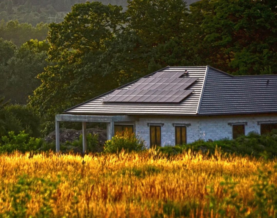 Wohnhaus mit dunkler Dachdeckung und Modulfläche, im Vordergrund eine Wiese im Abendlicht.